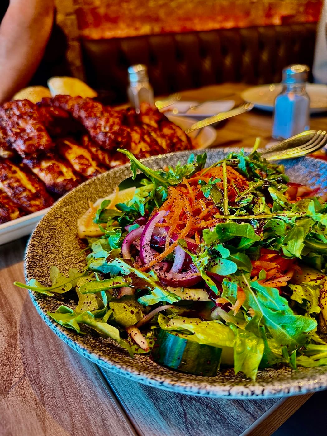 A bowl of fresh salad with rocket, red onion, cucumber and shredded carrot at Antep Kitchen Oxford, with the chargrilled platter behind it